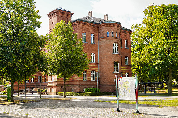 Ein historisches Backsteingebäude mit vielen Fenstern steht umgeben von Bäumen und Gras. Links ist ein Informationsschild über Nahverkehr und Umgebung zu sehen. Die Szene ist hell erleuchtet, was eine freundliche Atmosphäre vermittelt.