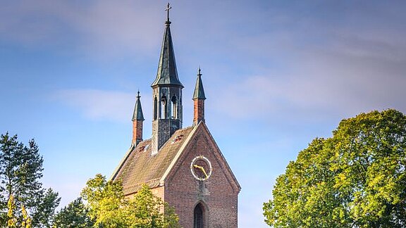 Ein historisches Gebäude mit einem hohen Turm und einer Uhr an der Fassade ist von grünen Bäumen umgeben. Der Himmel ist teilweise bewölkt und die Farben des Herbstes sind deutlich sichtbar.