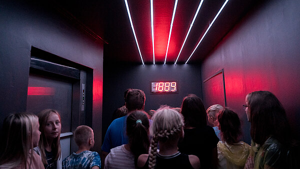 Eine Gruppe von Kindern steht in einem dunklen Flur mit rotem Licht. An der Wand hängt eine digitale Anzeige, die die Zahl "1889" zeigt. Der Raum hat eine futuristische Atmosphäre, die durch die Lichtstreifen an der Decke verstärkt wird.