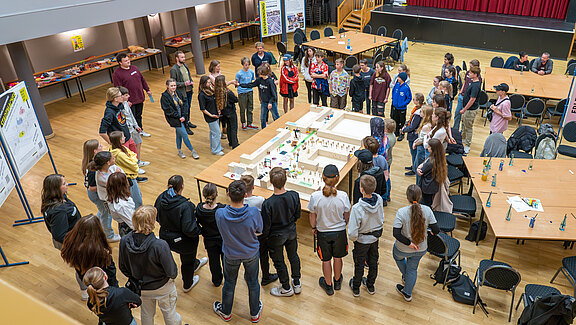 In einem Raum stehen etwa 30 Personen um einen großen Tisch, auf dem ein Modell angeordnet ist. Die Teilnehmer diskutieren und schauen aufmerksam auf das Modell. Die Atmosphäre wirkt interaktiv und gemeinschaftlich, mit verschiedenen Altersgruppen und Geschlechtern vertreten.