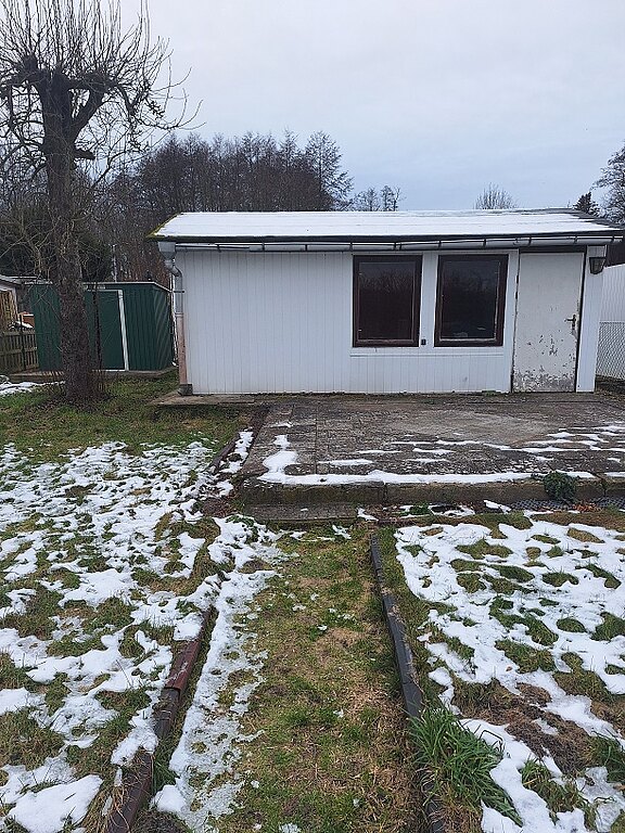 Ein einfacher, weißer Bungalow mit einem Flachdach steht in einem Garten, in dem schneeüberdeckter Boden und grüne Rasenteile sichtbar sind. Vor dem Gebäude verläuft ein gepflasterter Weg. Im Hintergrund sind Bäume und ein grünes Gartenhaus. Der Himmel ist bewölkt.