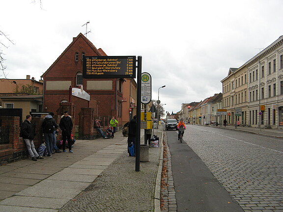 Bushaltestelle an städtischer Straße: Mehrere Menschen warten unter einem digitalen Anzeiger auf den Bus. Ein Radfahrer passiert auf Kopfsteinpflaster, Häuser säumen die Strecke.