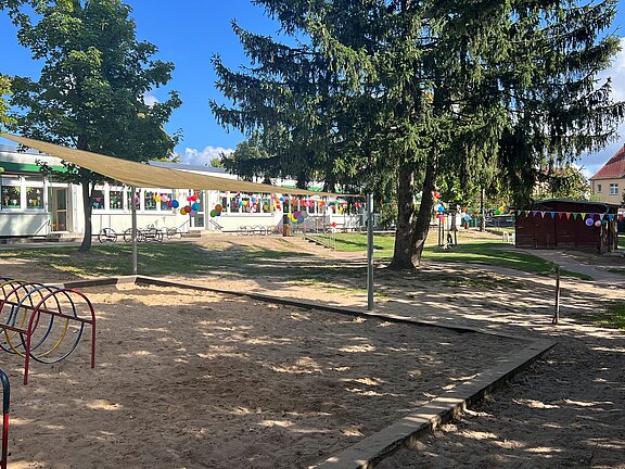 Der Blick auf einen Spielplatz mit einem Sandkasten und Klettergerüst im Vordergrund. Im Hintergrund steht ein mehrstöckiges Gebäude, umgeben von Bäumen und bunten Fähnchen, die festlich dekoriert sind. Der Himmel ist blau mit einigen Wolken.