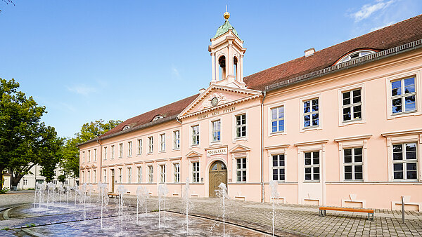 Das Bild zeigt ein historisches, rosafarbenes Gebäude mit einer Uhr und einem Turm. Vor dem Gebäude sprudeln mehrere Wasserfontänen auf einem gepflasterten Platz. Im Hintergrund sind Bäume zu sehen, und der Himmel ist klar und blau.