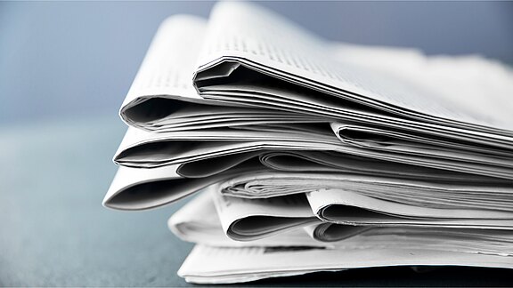 Ein Stapel von gefalteten Zeitungen liegt auf einem Tisch. Die Zeitungen sind übereinandergelegt und zeigen eine glänzende, weiße Oberfläche. Der Hintergrund ist unscharf und in einem sanften Blau gehalten.