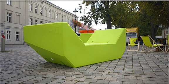 Eine große, leuchtend grüne Sitzgelegenheit in moderner, geometrischer Form steht auf einem Pflastersteinboden. Im Hintergrund sind einige Stühle und Gebäude sichtbar, die eine einladende Atmosphäre schaffen.
