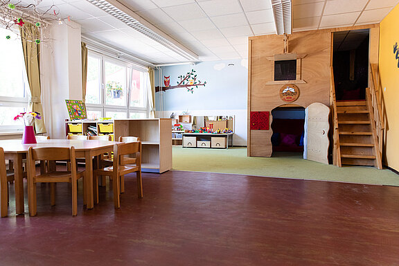 Der Raum ist ein helles Klassenzimmer mit einem großen Tisch aus Holz und Stühlen. An der Wand befindet sich ein Baum mit einer Eule. In der Ecke steht ein Spielbereich mit Kisten und Spielzeug. Rechts gibt es einen aufgebauten Spielplatz mit einer kleinen Treppe.