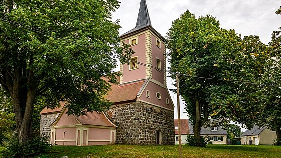Eine Steinkirche mit einem hohen, spitzen Turm und einem roten Satteldach. Die Fassade ist pinkfarben und von großen, grünen Bäumen umgeben. Im Hintergrund sind weitere Gebäude sichtbar. Der Himmel ist bewölkt.