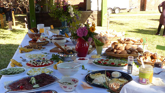 Ein reich gedeckter Tisch im Freien mit verschiedenen Speisen, darunter Salate, belegte Platten, Brötchen und ein bunter Blumenausfluss. Die Atmosphäre wirkt einladend und festlich, ideal für ein gemeinsames Essen im Freien.