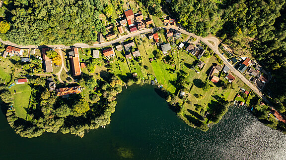 Luftaufnahme eines kleinen Dorfes am Wasser, umgeben von Bäumen. Die Häuser sind in einer Linie entlang einer Straße angeordnet, mit grünen Wiesen und Gärten. Der See reflektiert Sonnenlicht und ist von Pflanzen und Booten gesäumt.