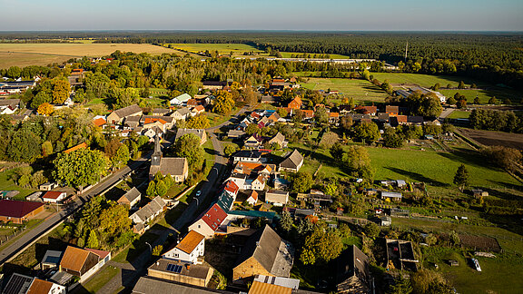 Luftaufnahme eines kleinen Dorfes umgeben von Wiesen und Feldern. Die Dächer der Häuser sind bunt und die Umgebung ist grün mit zahlreichen Bäumen. Im Hintergrund erstreckt sich ein Wald, während die Landschaft weitläufig und idyllisch wirkt. Ein klarer blauer Himmel ist sichtbar.