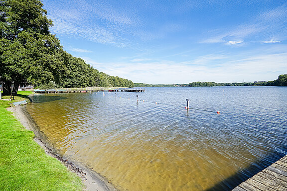 Ein ruhiger See mit klarem Wasser und einer grünen Uferlandschaft. Am Rand stehen Bäume, während am Wasser ein Steg sichtbar ist. Die Himmel ist blau mit einigen Wolken. Am Wasser sind Absperrungen aus Bojen zu sehen.