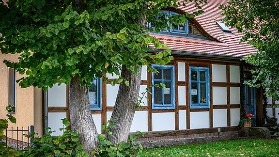 Das Bild zeigt ein traditionelles Haus mit Fachwerk und blauen Fenstern. Das Dach ist rot, und der Bereich vor dem Haus ist von einem gepflegten Rasen und grünen Bäumen umgeben. Ein ruhiger, ländlicher Eindruck wird vermittelt.