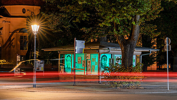 Eine beleuchtete Bushaltestelle in der Nacht, umgeben von Bäumen. An den Fenstern sind Kunstwerke und Porträts sichtbar. Eine Straßenlaterne erzeugt warmes Licht, während Auto- und Stadtlichter im Hintergrund schemenhaft zu sehen sind.