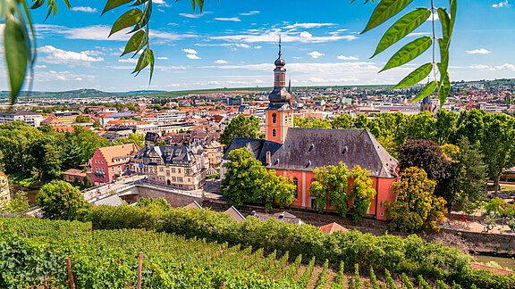 Ein malerischer Blick auf die Stadt, umgeben von Weinbergen. Im Vordergrund ist eine große, orangefarbene Kirche mit einer hohen Spitze, während sich dahinter eine Vielzahl von Gebäuden und grünen Bäumen erstreckt. Der Himmel ist blau mit einigen Wolken.