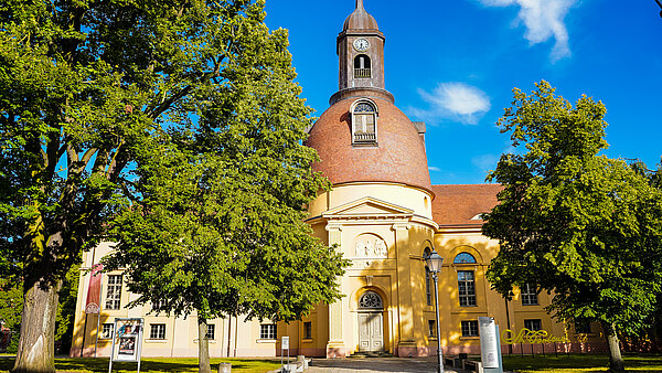 Eine gelbe Kirche mit einer großen, runden Kuppel und einem Glockenturm steht inmitten von grünen Bäumen. Der Himmel ist blau mit einigen Wolken. Die Fassade der Kirche zeigt architektonische Details, einschließlich einer Haupttür und Fenstern.