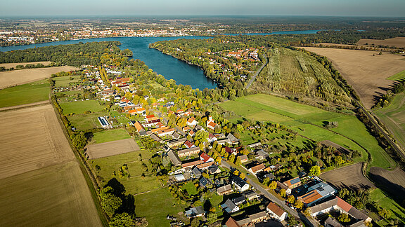 Eine Luftaufnahme eines friedlichen ländlichen Gebiets, das von Feldern, Wohnhäusern und einem Fluss umgeben ist. Das Bild zeigt dichte grüne Bäume entlang des Flussufers und eine kleine Ortschaft mit bunten Dächern im Herbstlicht.