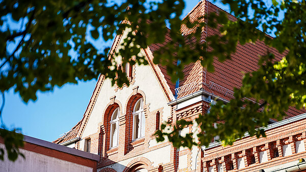 Das Bild zeigt die Dachansicht eines ehemaligen Gebäudes mit roten Ziegeln und dekorativen Elementen. Auffällig sind die großen, runden Fenster im oberen Bereich und die spitzen Dächer, die unter einem klaren blauen Himmel sichtbar sind. 일부 소나무가 이미지 일부를 가리고 있습니다.