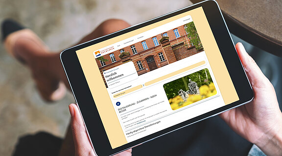 Nahaufnahme eines Tablets in Händen: Auf dem Bildschirm ist die Startseite einer städtischen Verwaltungs-Website mit Begrüßung, Foto eines Backstein-Gebäudes und Navigationsbereichen zu Dienstleistungen zu sehen.