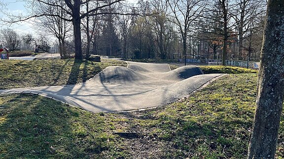 Eine Sicht auf einen Skatepark mit einer geschwungenen Betonbahn, die sanfte Hügel und Mulden bildet. Die Umgebung ist grün mit Gras und einigen Bäumen. Die Szene ist hell erleuchtet, und es gibt keine Personen im Bild.