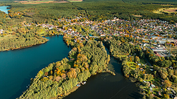 Luftaufnahme eines malerischen Dorfes, das an einem Fluss und Seen liegt. Umgeben von Wäldern und buntem Herbstlaub zeigt die Landschaft eine ruhige, natürliche Umgebung mit Wasserflächen und grünen Uferzonen. Einige Boote sind am Rand zu sehen.