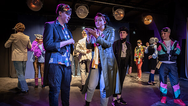 Die Bühne zeigt eine Gruppe von Schauspielern in verschiedenen Kostümen, die miteinander interagieren. Eine Person in einem langen Mantel spricht mit einem anderen, während im Hintergrund verschiedene Figuren in farbenfrohen Outfits stehen, die die dynamische Atmosphäre einer Theateraufführung vermitteln.