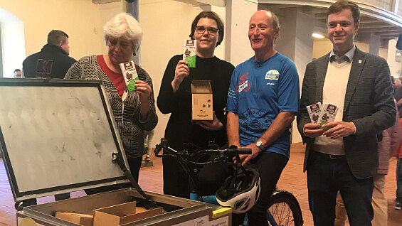 Die Abbildung zeigt vier Personen, die an einem Fahrrad mit einem offenen Kasten stehen. Die Personen halten verschiedene Informationsmaterialien zur Klimainitiative Schmöckwitz in den Händen. Im Hintergrund ist ein heller Raum zu erkennen.