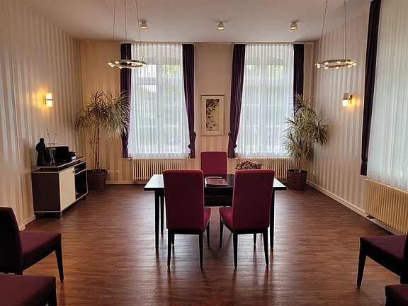 Ein heller Raum mit Holzfußboden und großen Fenstern, die von lila Vorhängen umrahmt sind. In der Mitte stehen ein Tisch und vier Stühle. An den Wänden stehen Pflanzen. Auf einem Tisch in der Ecke befindet sich ein kleiner schwarzer Schrank.