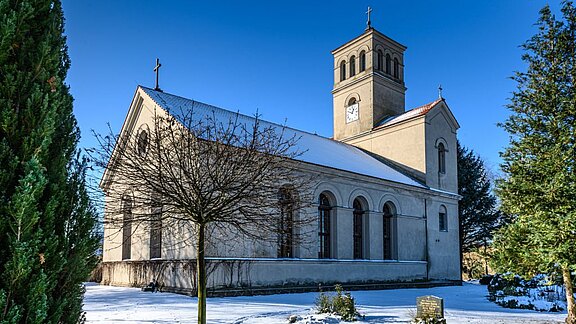 Eine schlichte Kirche mit einem Glockenturm steht in einer winterlichen Landschaft. Der Boden ist mit Schnee bedeckt, und ein kahler Baum steht in der Nähe. Im Hintergrund sieht man Nadelbäume und einen klaren blauen Himmel.
