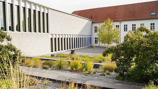 Im Fokus der Szene steht ein moderner Gebäudekomplex mit großen Fenstern, umgeben von einem angelegten Garten mit bunten Pflanzen und schmalen Wegen. Ein grüner Baum und eine kleine Statuette sind ebenfalls sichtbar. Der Garten vermittelt eine ruhige und einladende Atmosphäre.