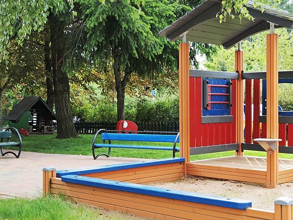 Ein Spielplatz mit einer hölzernen Spielstruktur, die bunt gestaltet ist, sowie einer Sandfläche. Im Hintergrund stehen Bäume und eine grüne Wiese. Zwei Bänke sind in der Nähe aufgestellt, und ein Spielhaus ist ebenfalls zu sehen.