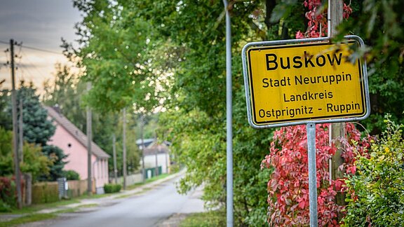 Das Bild zeigt ein Ortsschild mit der Aufschrift „Buskow, Stadt Neuruppin, Landkreis Ostprignitz-Ruppin“. Es befindet sich am Straßenrand, umgeben von Bäumen und Pflanzen. Im Hintergrund ist eine Straße sichtbar, die in ein Wohngebiet führt.