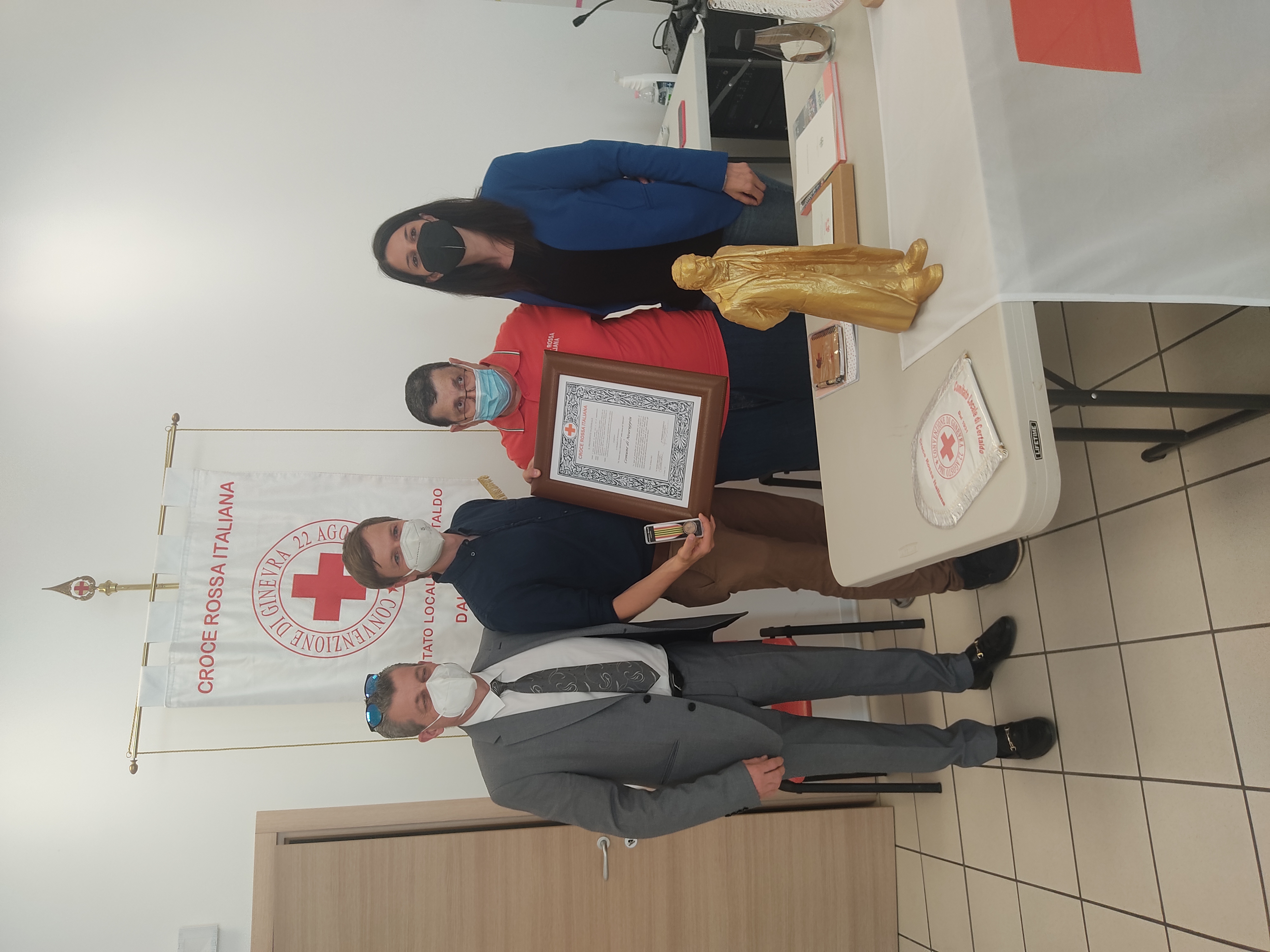 Vier Personen stehen an einem Tisch, der mit der Flagge des Roten Kreuzes dekoriert ist. Sie halten eine Urkunde und posieren zusammen. Im Hintergrund ist ein Banner des Roten Kreuzes sichtbar. Auf dem Tisch stehen verschiedene Gegenstände, darunter eine goldene Statue.