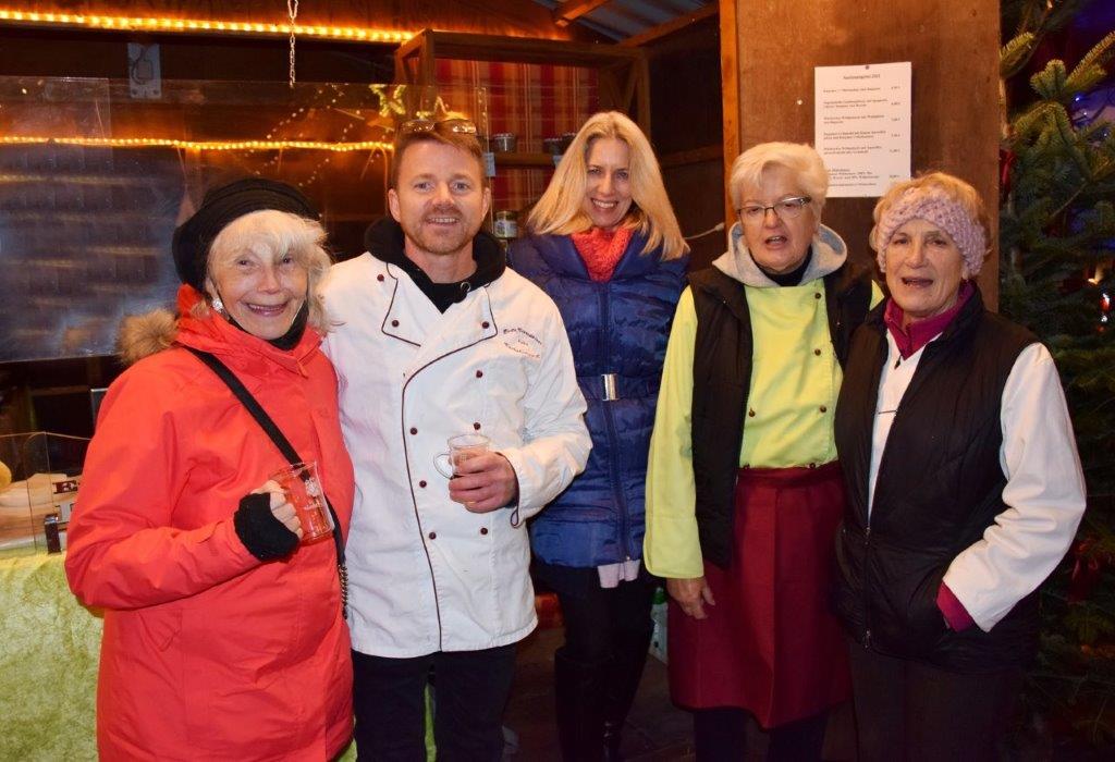 Eine Gruppe von fünf Personen steht zusammen in einem festlichen Umfeld. Sie lächeln und tragen warme Winterkleidung. Ein Mann in einer Kochjacke ist in der Mitte, umgeben von Frauen in unterschiedlichen Jacken und Mützen. Es herrscht eine fröhliche Stimmung.