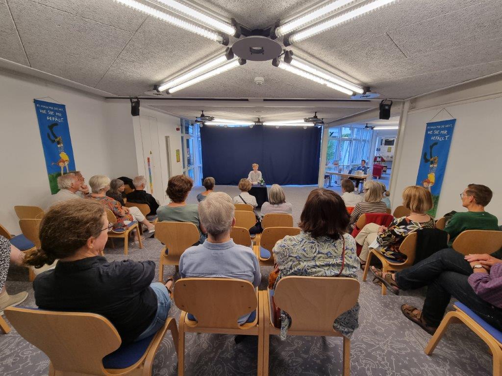 Eine Gruppe von Menschen sitzt in einem Raum auf Stühlen und schaut auf eine Bühne, auf der eine Person spricht. Der Raum hat eine helle Beleuchtung, und im Hintergrund sind zwei Banner sichtbar. Die Zuschauer scheinen interessiert zuzuhören.