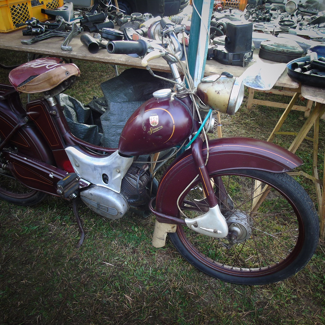 Ein antikes Motorrad in rot-brauner Farbe steht auf einer Wiese. Es hat einen klassischen Look mit einer runden Frontscheinwerfer, einem bequemen Sitz und technischen Teilen im Hintergrund, die auf einem Tisch ausgelegt sind.