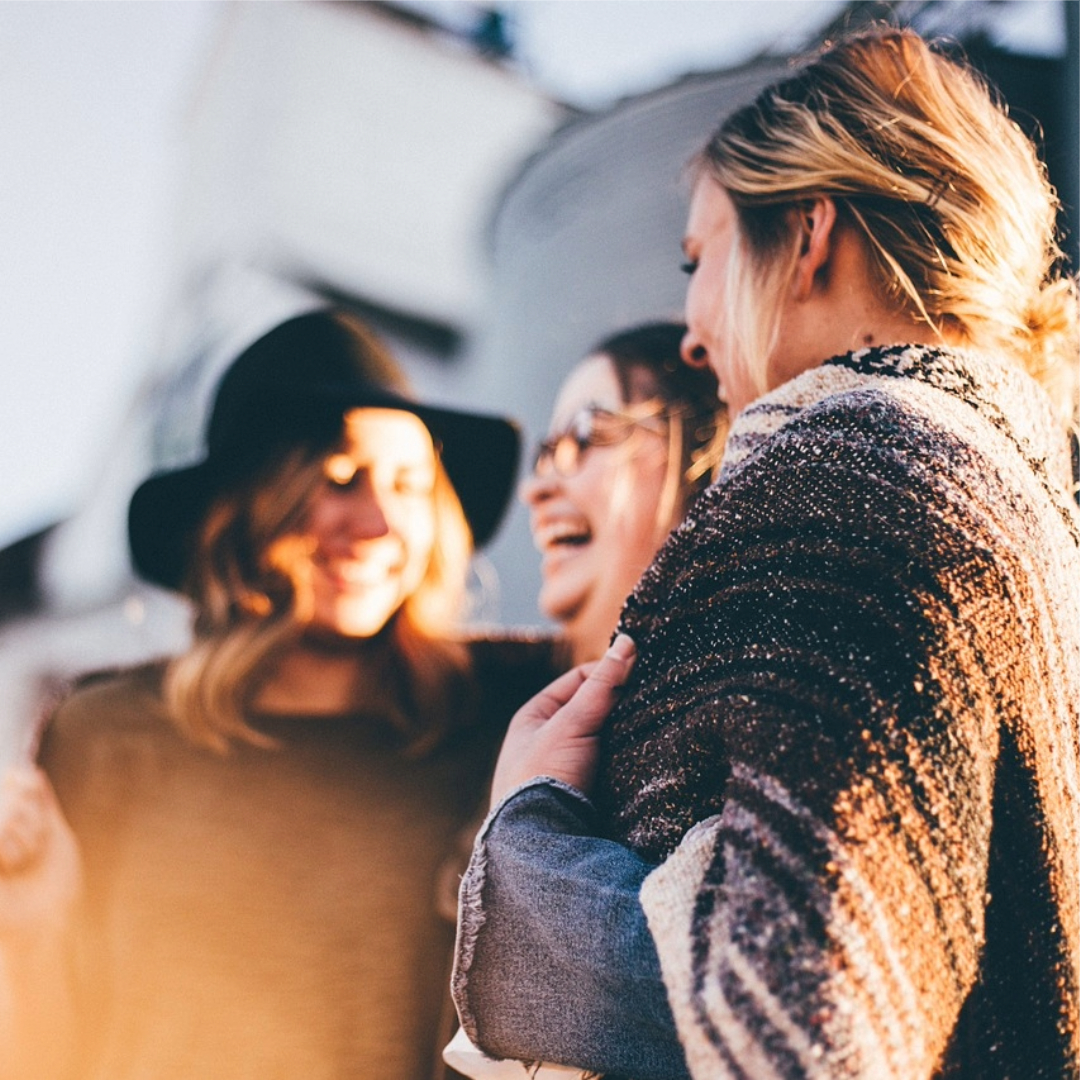 Eine Gruppe von drei lächelnden Frauen, die miteinander sprechen und lachen. Die Frauen tragen verschiedene Kleidung und scheinen eine fröhliche Zeit im Freien zu haben. Die Umgebung wirkt freundlich und einladend, mit sanften Farben und diffusem Licht.