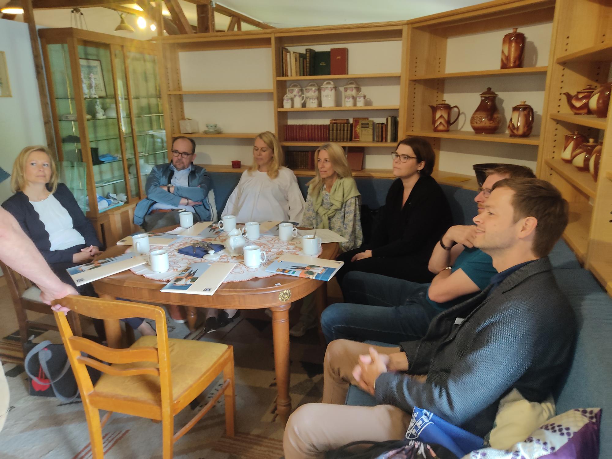 Eine Gruppe von sieben Personen sitzt um einen Tisch in einem hellen Raum. Einige halten Tassen, während sie aufmerksam miteinander sprechen. In der Mitte des Tisches liegen Flyer und Materialien. Der Raum ist gemütlich und bietet Regale mit Büchern und Töpfen im Hintergrund.