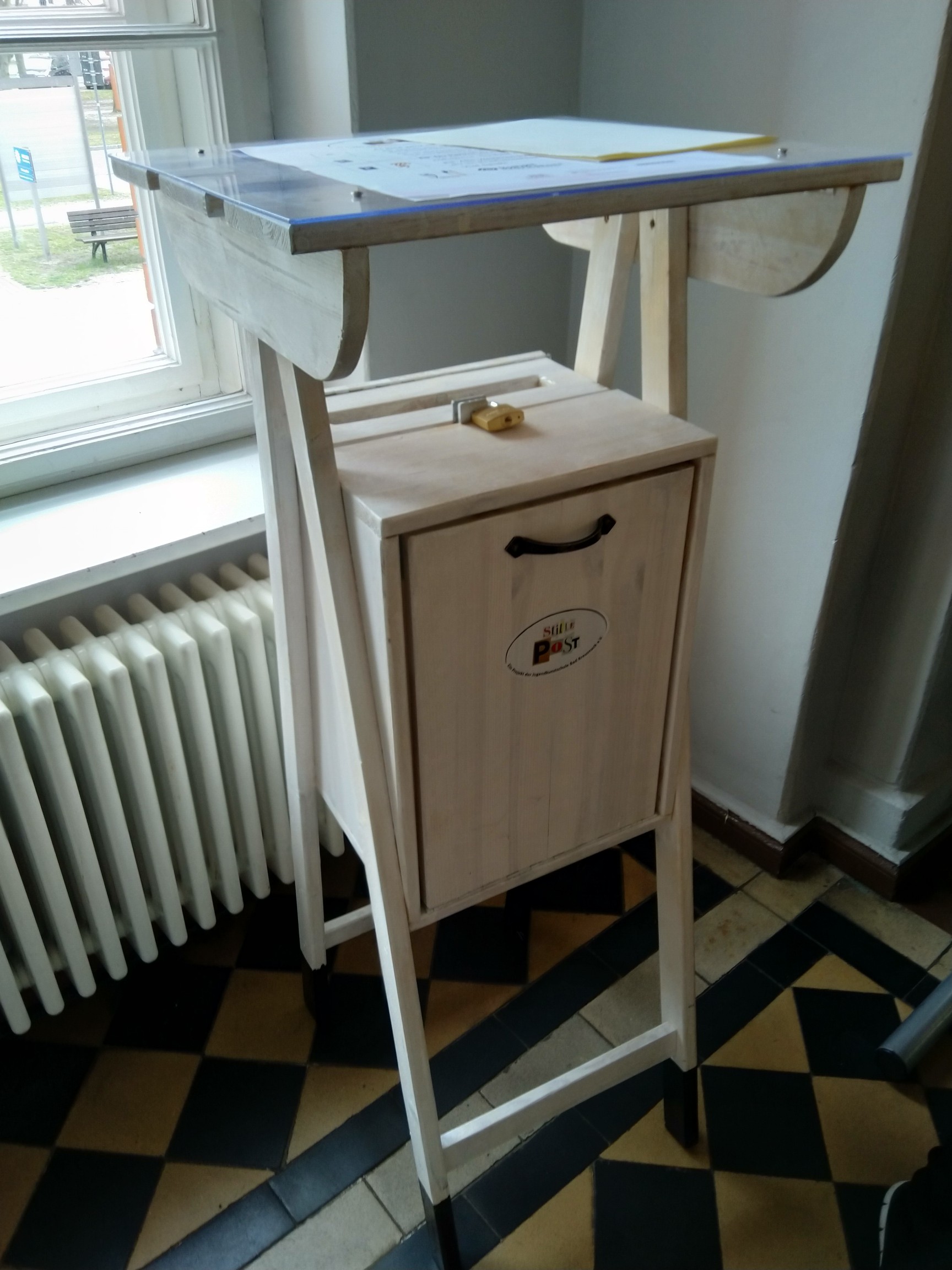 Hohe hölzerne Sammelbox mit integriertem Stehpult. Oben eine flache Schreibfläche mit Dokument unter Plexi; darunter ein verschließbarer Kasten mit Griff und Etikett „Post“. Dient zum Einwerfen von Nachrichten oder Vorschlägen.