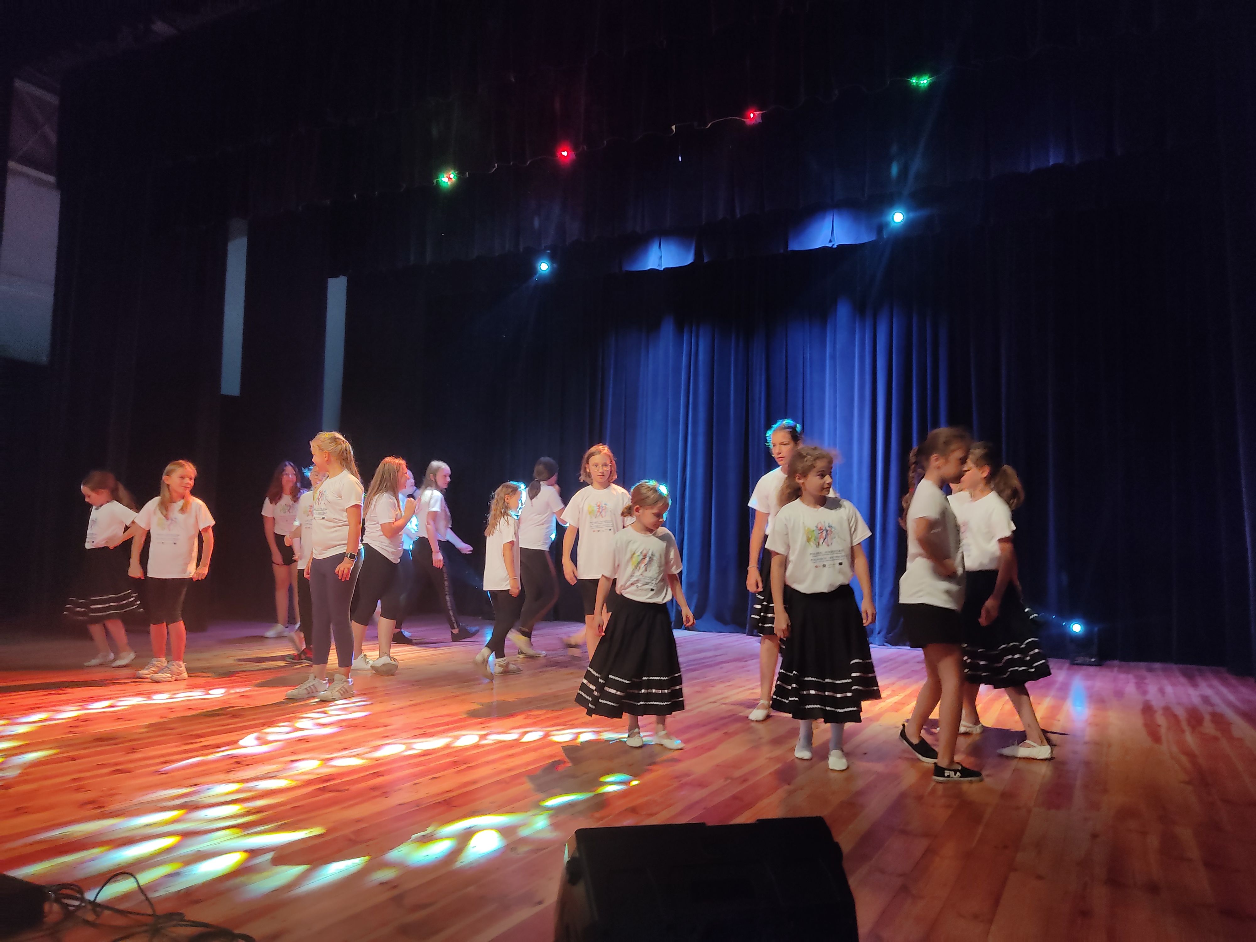 Eine Gruppe Kinder und Jugendlicher tanzt auf einer Theaterbühne. Sie tragen weiße T‑Shirts; einige Mädchen schwarze Röcke. Bunte Scheinwerfer beleuchten den Holzboden, hinter ihnen ein dunkelblauer Vorhang. Gruppenchoreografie in Aufführung.