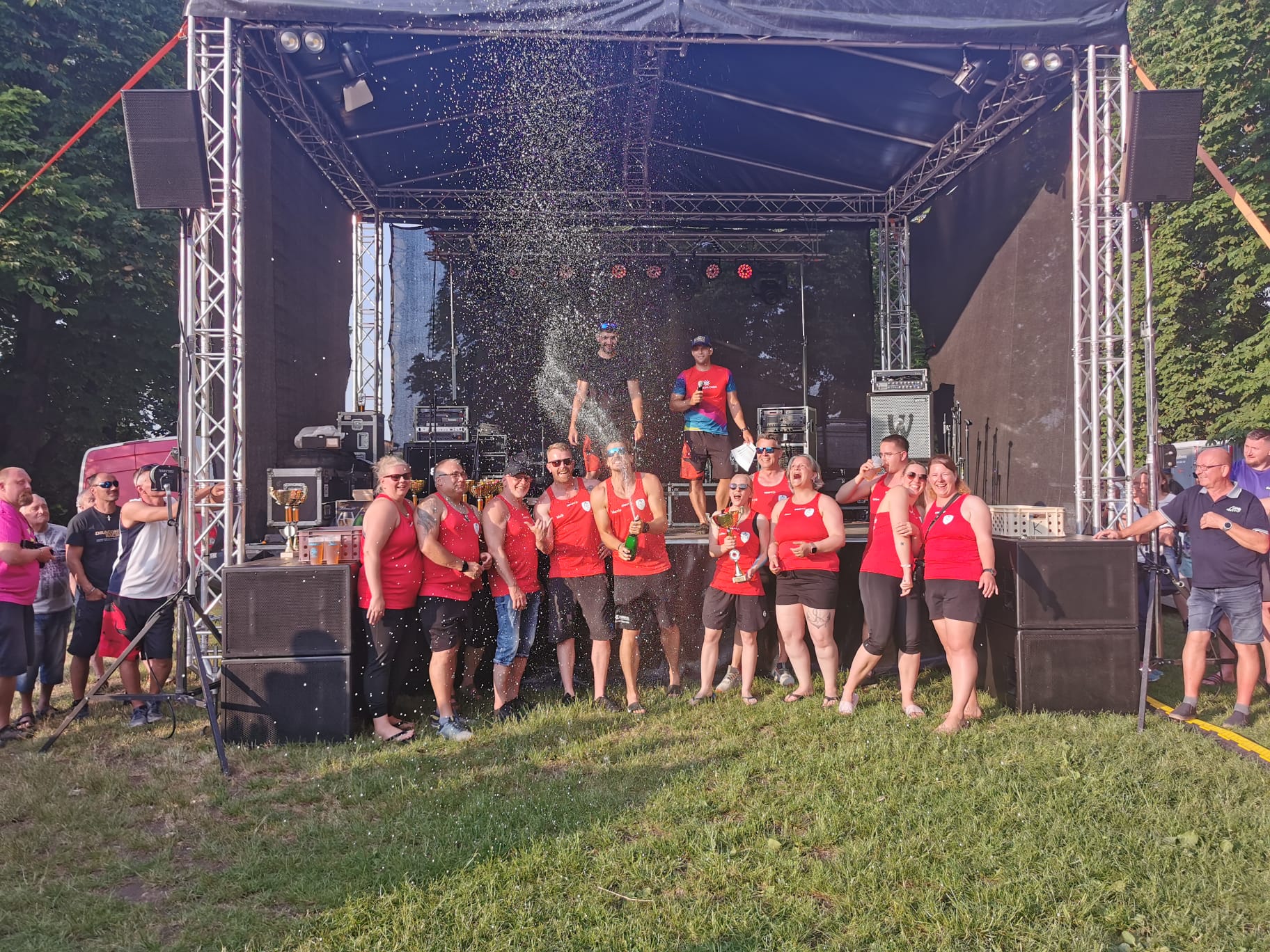 Gruppe in roten Trikots feiert vor einer Open‑Air‑Bühne. Mehrere tragen Medaillen, jemand hält einen Pokal, Sekt spritzt in die Luft. Auf der Bühne stehen weitere Personen mit Trophäen. Publikum beobachtet. Stimmung: Siegesfeier nach Wettkampf.