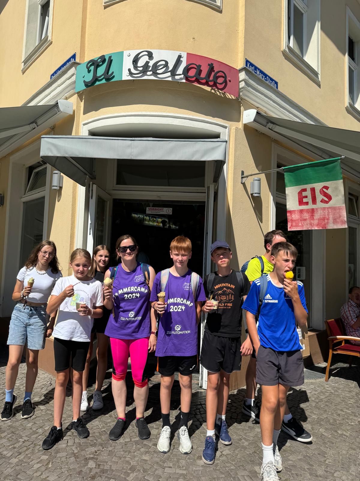 Eine Gruppe Jugendlicher steht vor einer Eisdiele und isst Eis. Sie lachen, halten Becher und Waffeln, sommerliche Kleidung. Über dem Eingang steht „Il Gelato“, daneben ein „EIS“-Schild. Sonniges Wetter, entspannte Stimmung.