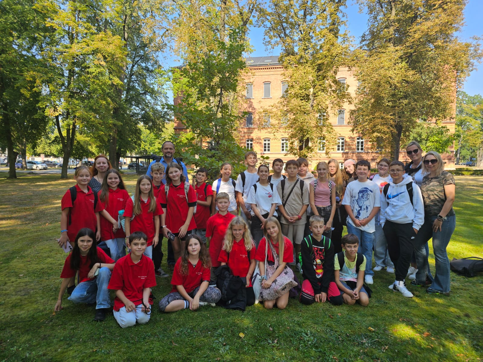 Gruppenfoto einer Schulklasse im Freien. Rund dreißig Kinder und mehrere Erwachsene posieren auf einer Wiese, viele Kinder tragen rote T-Shirts. Hinter ihnen stehen Bäume, im Hintergrund ein Backsteingebäude. Sonniger Tag, fröhliche Stimmung.