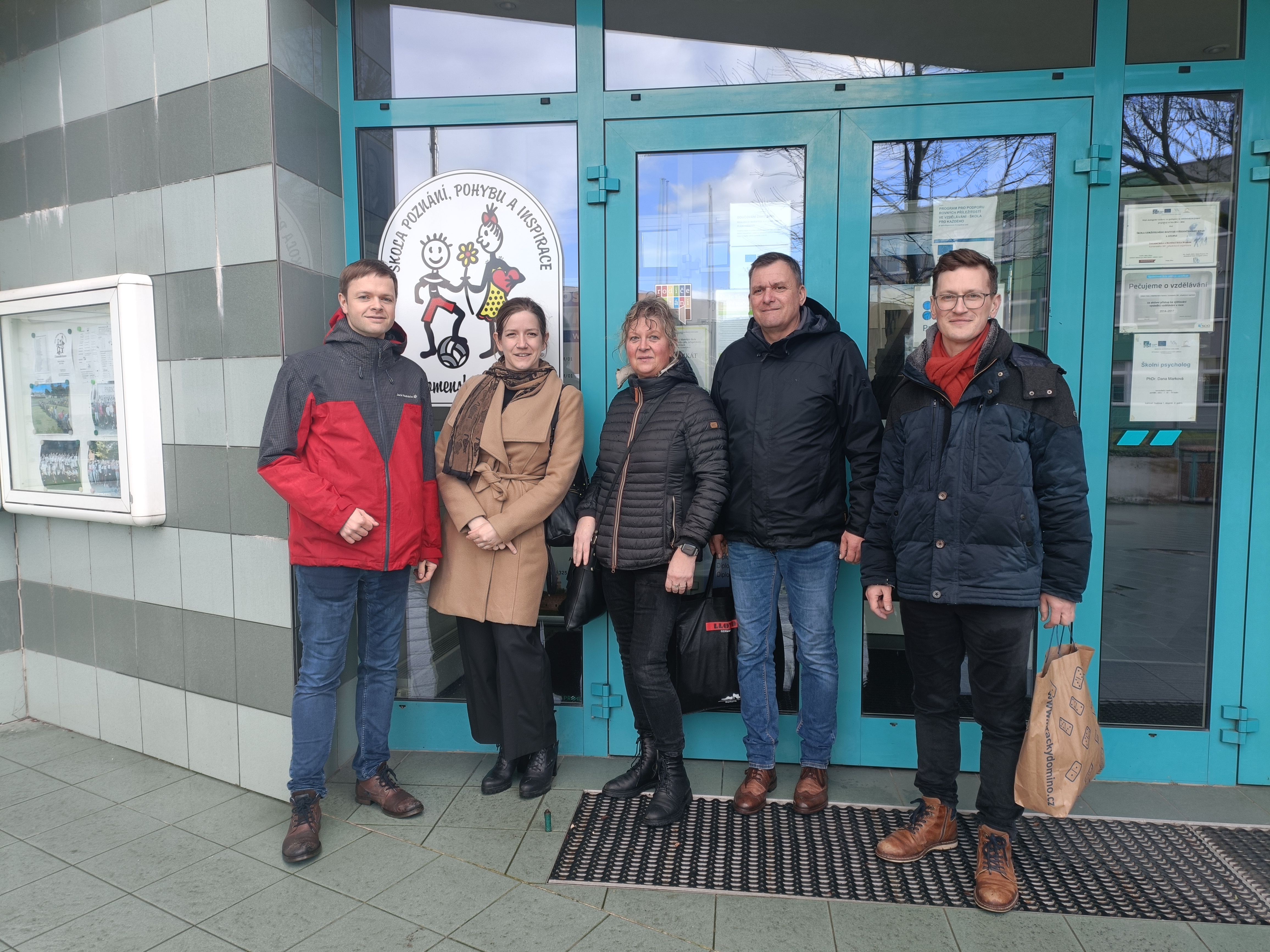 Fünf Erwachsene stehen lächelnd vor einem Gebäudeeingang mit türkisfarbenen Türen. Neben ihnen hängt ein rundes Logo mit Comic-Kindern, das auf eine Bildungseinrichtung hindeutet. Gruppenfoto bei kühlem Wetter, alle tragen Jacken.