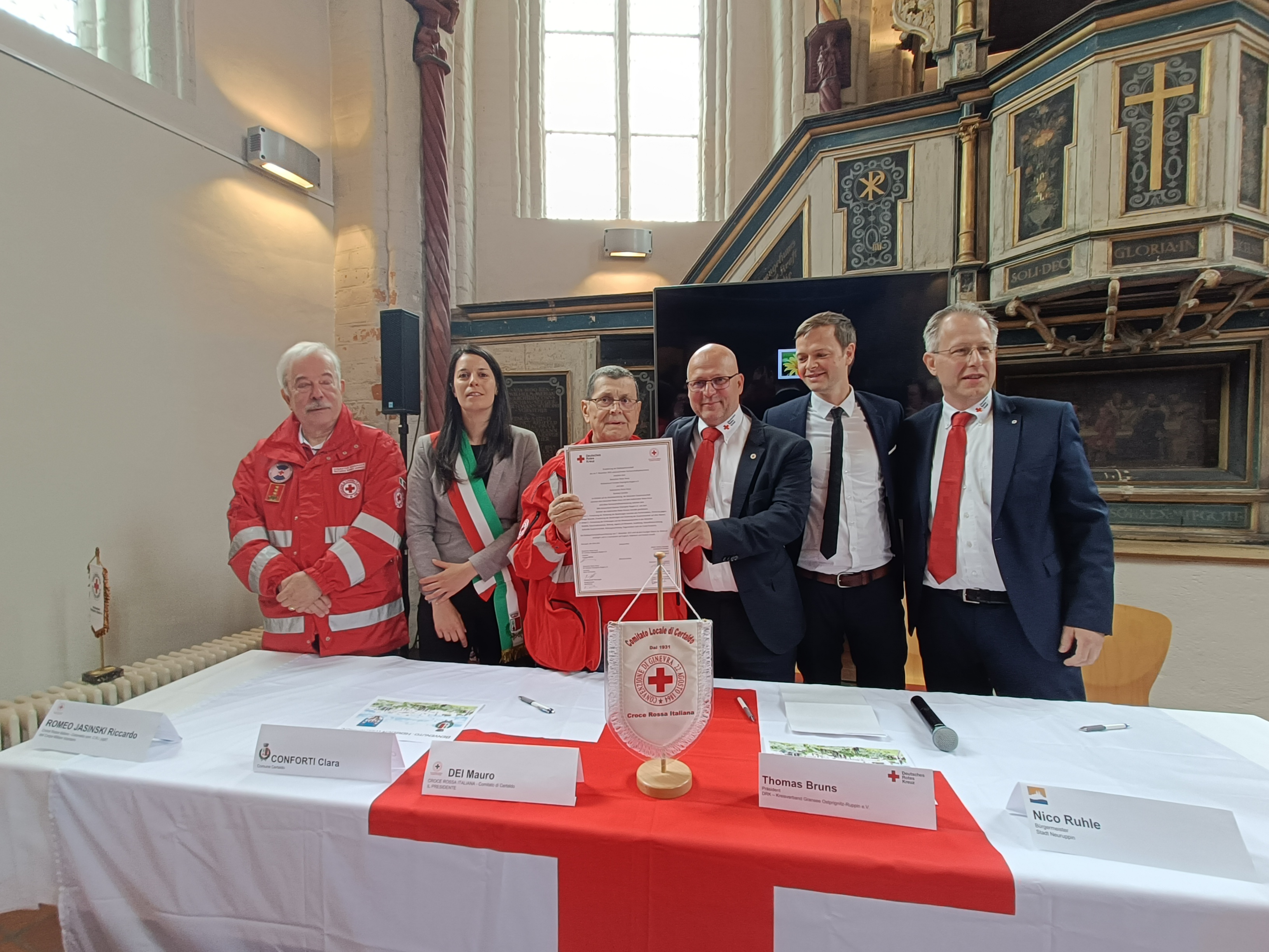 Sechs Personen, teils in Rotkreuz-Uniform, stehen hinter einem Tisch mit Rotkreuz-Flagge und Emblem. In der Mitte wird ein unterschriebenes Dokument hochgehalten – eine feierliche Unterzeichnung oder Partnerschaft in einer Kirchenhalle.