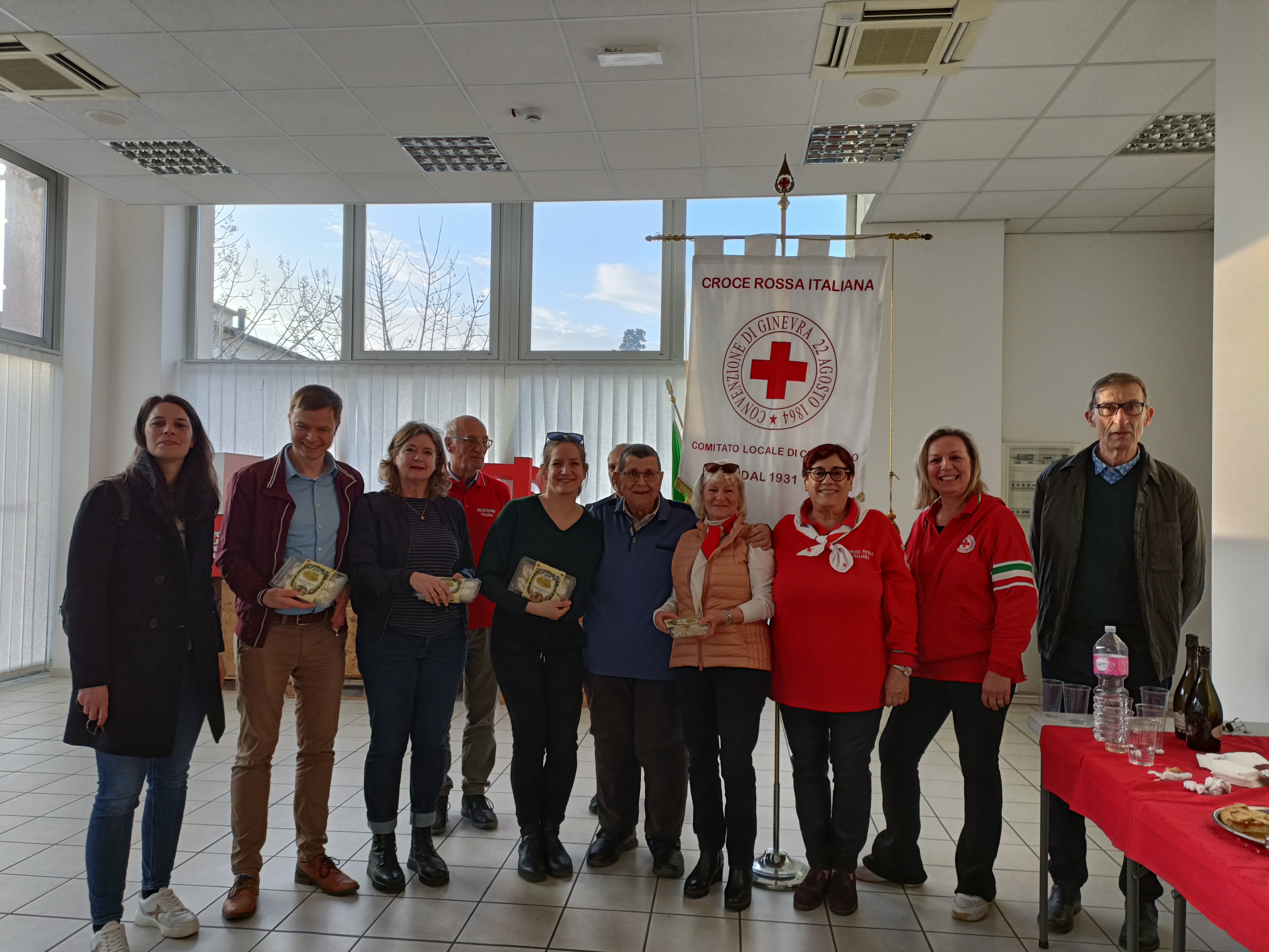 Gruppe von Freiwilligen steht in einem hellen Raum vor einem Banner der Croce Rossa Italiana. Mehrere halten Auszeichnungen in den Händen. Die Szene wirkt wie eine kleine Ehrung oder Dankesfeier.