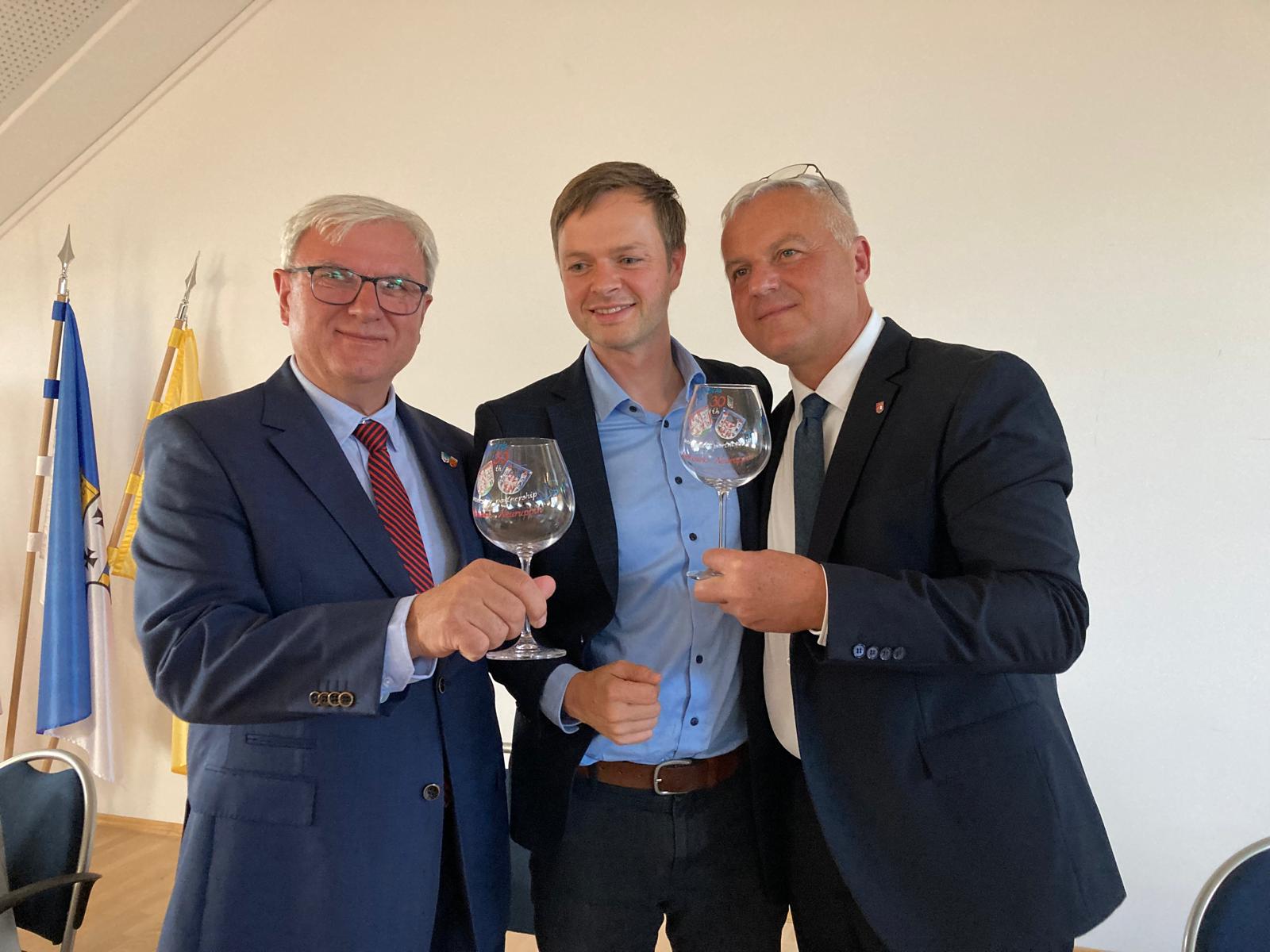 Drei Männer im Anzug stoßen mit Weingläsern an und lächeln. Sie stehen eng beieinander in einem hellen Raum; im Hintergrund sind Fahnen sichtbar.