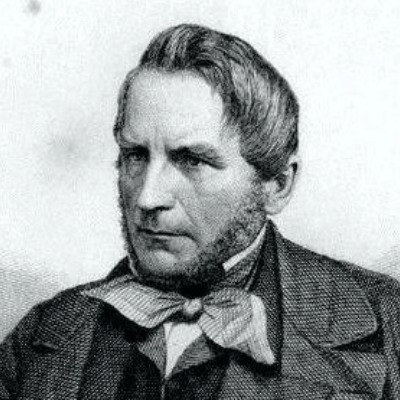 Porträt eines Mannes mit einem ernsten Ausdruck, kurzen, grauen Haaren und einem Bart. Er trägt einen Anzug mit einer Krawatte und eine Schärpe um den Hals. Der Hintergrund ist unscharf, um den Fokus auf das Gesicht zu lenken.