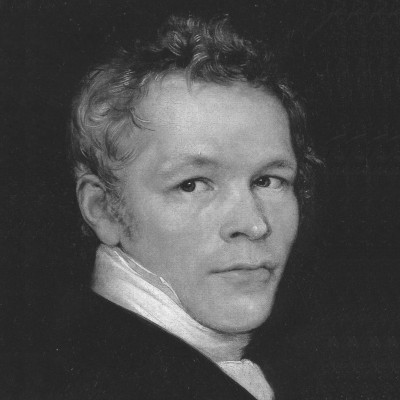 Porträt eines Mannes mit kurzem, lockigem Haar und einem nachdenklichen Gesichtsausdruck. Er trägt einen dunklen Anzug und hat einen weißen Kragen. Das Bild hat einen schlichten, dunklen Hintergrund, der die Gesichtszüge betont.