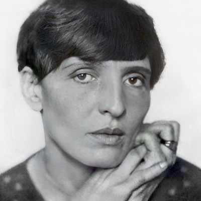 Ein Porträt einer Frau mit kurzem, dunklem Haar, die direkt in die Kamera schaut. Sie hat einen nachdenklichen Gesichtsausdruck, während sie eine Hand an ihr Kinn hält. Der Hintergrund ist neutral, wodurch der Fokus auf ihrem Gesicht liegt.